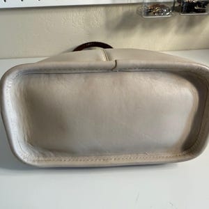 May include: A light beige leather handbag with a rounded rectangular base. The bag has a dark brown handle and a stitched seam detail around the bottom edge. The leather appears to be slightly worn.