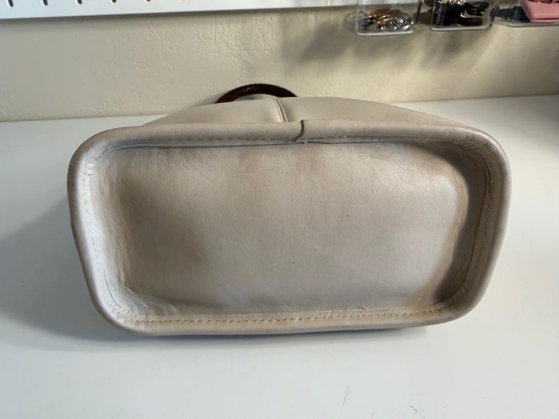 May include: A light beige leather handbag with a rounded rectangular base. The bag has a dark brown handle and a stitched seam detail around the bottom edge. The leather appears to be slightly worn.