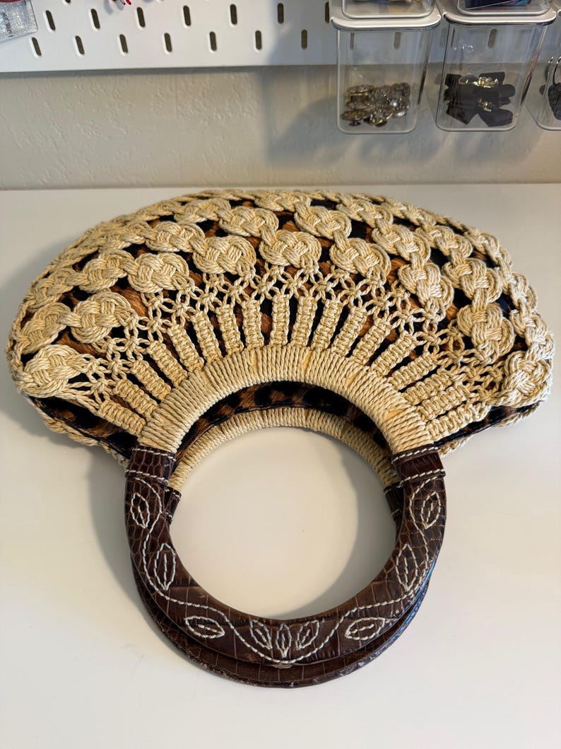 Op de afbeelding: Een beige en bruine handtas met een ronde handgreep. De tas heeft een geweven ontwerp met een luipaardprint voering. De handgreep is bruin met witte stiksels en een bladerenpatroon.