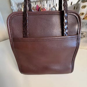 May include: A brown leather handbag with braided handles. The bag has a rectangular shape with a front pocket and a stitched border. The handles are dark brown and appear to be made of leather as well. The bag is a solid color.