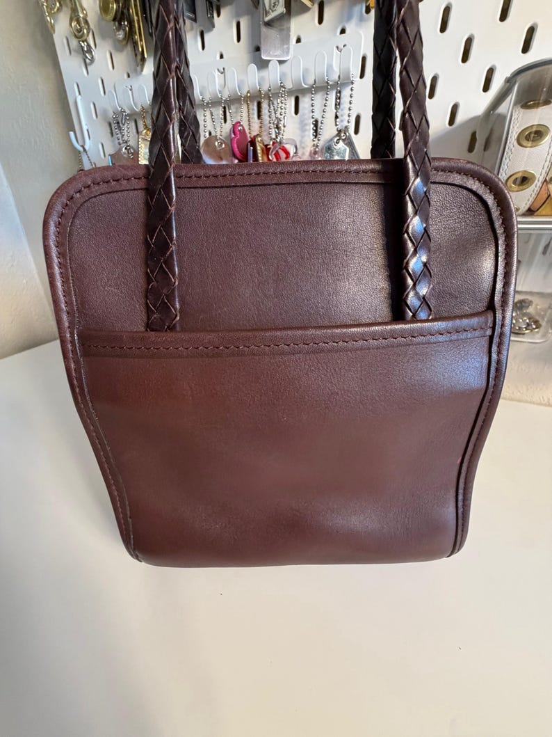 May include: A brown leather handbag with braided handles. The bag has a rectangular shape with a front pocket and a stitched border. The handles are dark brown and appear to be made of leather as well. The bag is a solid color.