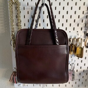 May include: A dark brown leather handbag with braided handles. The square-shaped bag has a front pocket and is hanging on a white pegboard. Gold-colored jewelry hangs on the pegboard to the left and right of the bag.