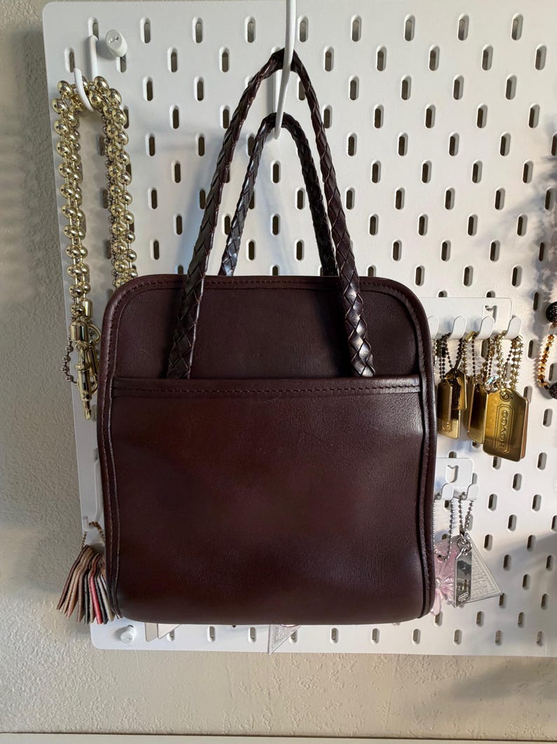 May include: A dark brown leather handbag with braided handles. The square-shaped bag has a front pocket and is hanging on a white pegboard. Gold-colored jewelry hangs on the pegboard to the left and right of the bag.
