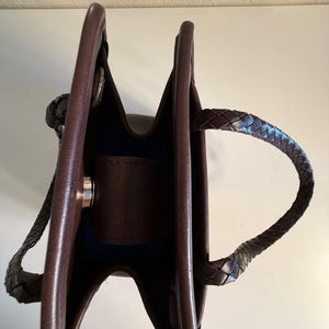 May include: A brown leather tote bag with braided handles. The bag has a structured design with a dark interior and a silver-colored clasp. The handles are securely attached to the bag's body, and the leather appears to be of high quality.
