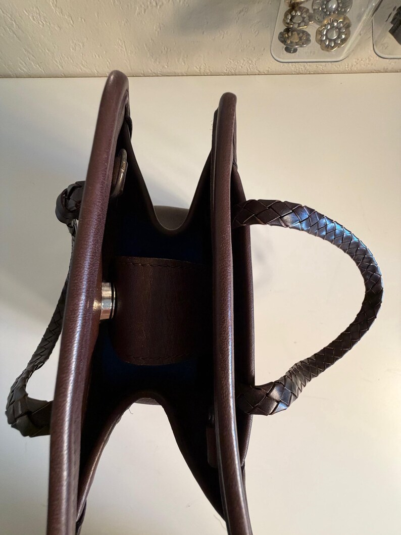 May include: A brown leather tote bag with braided handles. The bag has a structured design with a dark interior and a silver-colored clasp. The handles are securely attached to the bag's body, and the leather appears to be of high quality.