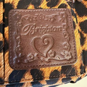 Op de afbeelding: Close-up van een bruin leren label met het woord "Brighton" in reli&euml;f en een hartontwerp, tegen een luipaardprintstof. De patch is rechthoekig met bloemen- en werveldetails langs de randen. Een rits is zichtbaar aan de bovenkant.