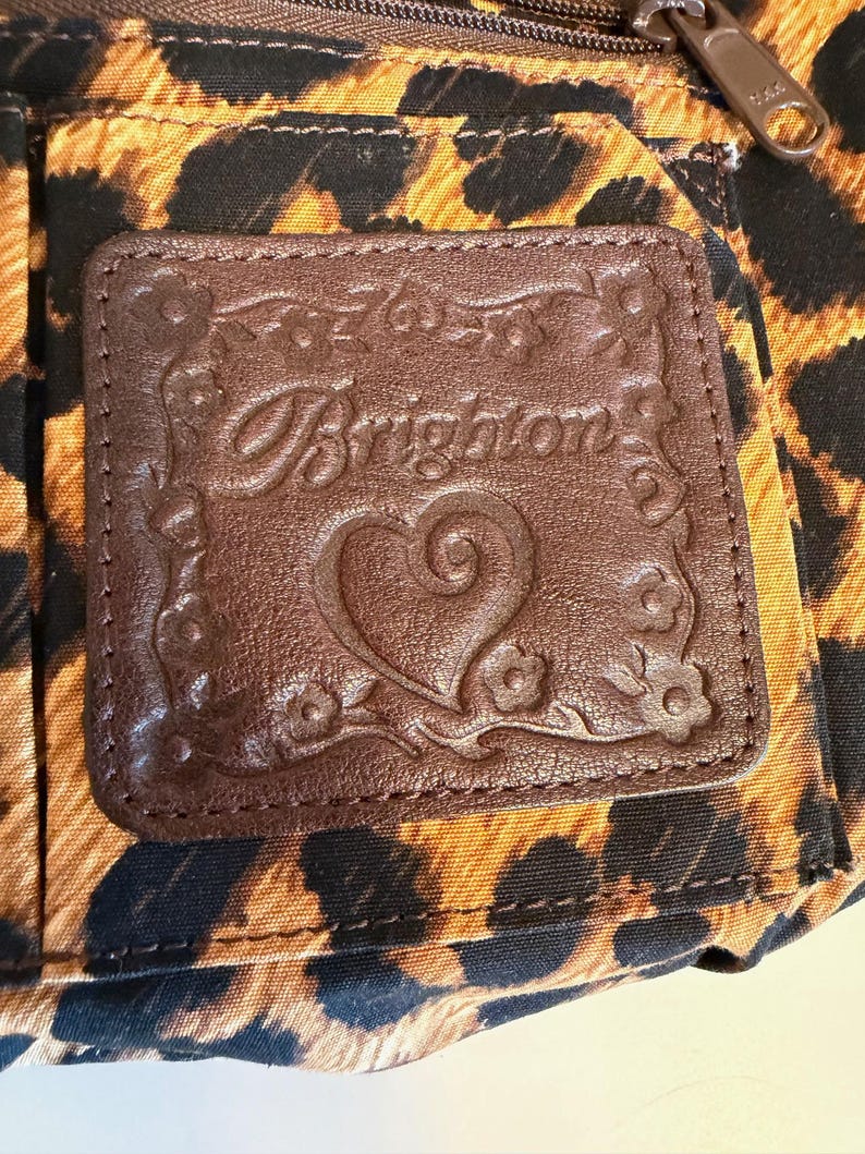 Op de afbeelding: Close-up van een bruin leren label met het woord "Brighton" in reli&euml;f en een hartontwerp, tegen een luipaardprintstof. De patch is rechthoekig met bloemen- en werveldetails langs de randen. Een rits is zichtbaar aan de bovenkant.