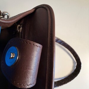 May include: Close-up of a brown leather handbag with a braided handle. The bag features a magnetic snap closure with a blue accent. The zipper is visible, and the leather appears to be smooth and well-maintained.