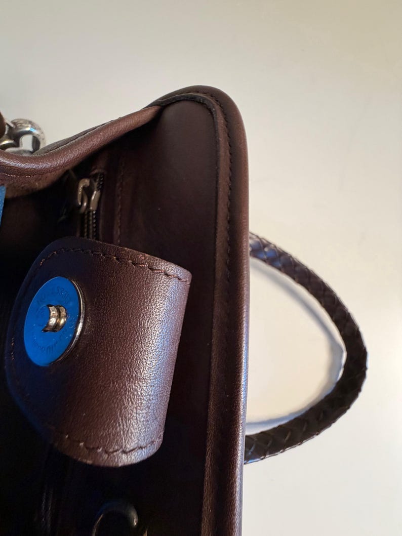 May include: Close-up of a brown leather handbag with a braided handle. The bag features a magnetic snap closure with a blue accent. The zipper is visible, and the leather appears to be smooth and well-maintained.
