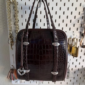 May include: A dark brown, square-shaped handbag with a crocodile skin texture. The bag has braided handles and silver-toned hardware. A heart-shaped charm hangs from the front. The bag is displayed against a white pegboard.