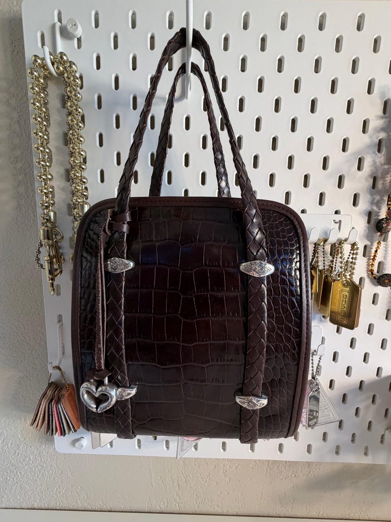 May include: A dark brown, square-shaped handbag with a crocodile skin texture. The bag has braided handles and silver-toned hardware. A heart-shaped charm hangs from the front. The bag is displayed against a white pegboard.