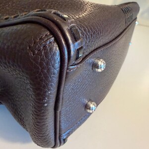 May include: Close-up of a dark brown leather handbag. The bag has a textured surface, decorative stitching, and silver-toned metal feet. The corners are reinforced with additional leather and stitching.