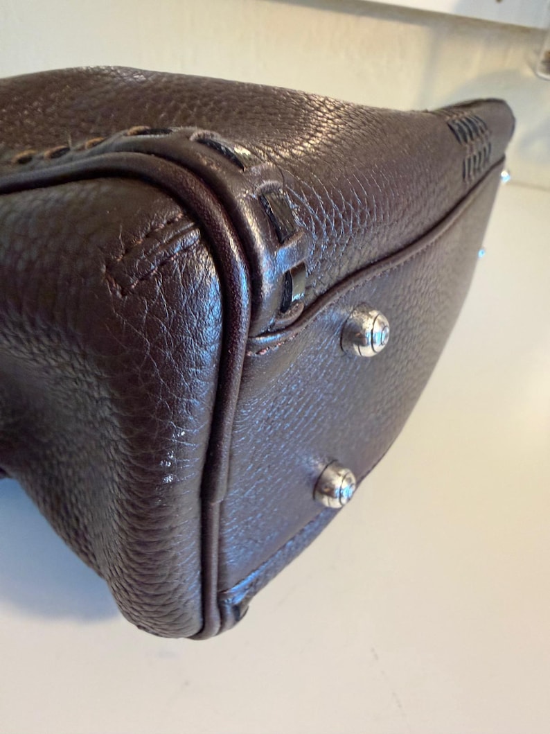 May include: Close-up of a dark brown leather handbag. The bag has a textured surface, decorative stitching, and silver-toned metal feet. The corners are reinforced with additional leather and stitching.