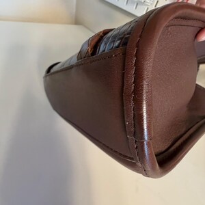 May include: A brown leather handbag with a woven top section. The bag has a rectangular shape with rounded corners and a stitched seam detail. The leather appears to be a rich, dark brown color.