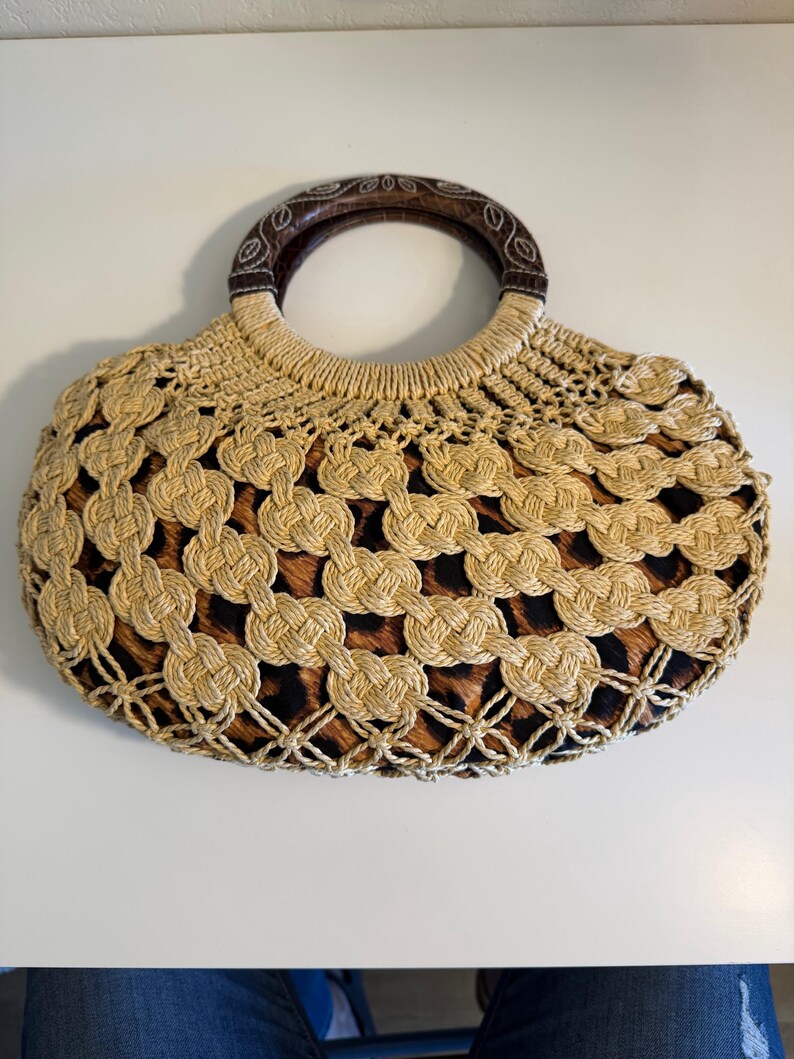 Op de afbeelding: Een beige geweven handtas met een bruin luipaardprint interieur. De tas heeft een ronde houten handgreep en een geweven ontwerp. Een stijlvol accessoire voor het dragen van essentials.