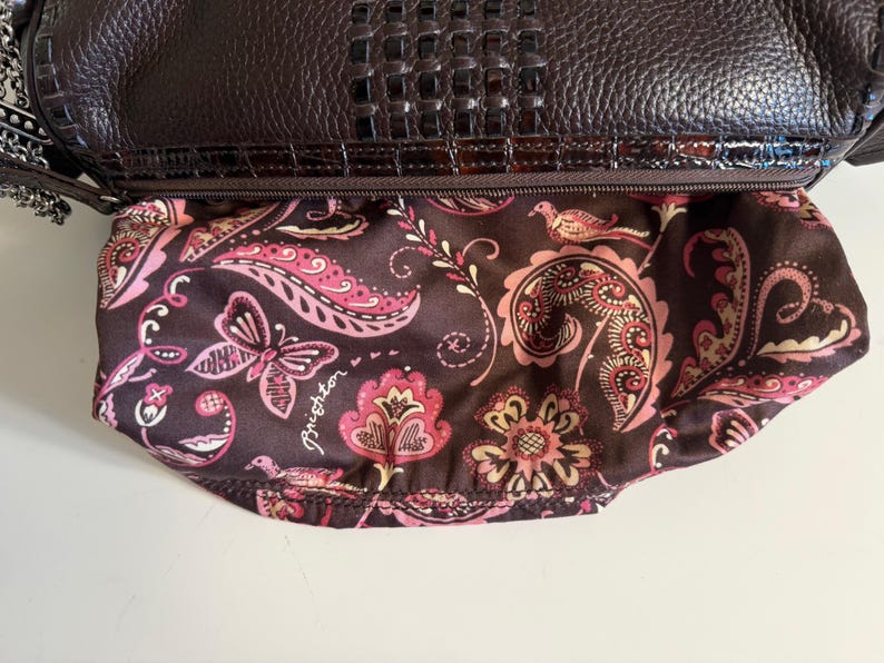 May include: A dark brown leather handbag with a textured top flap and a patterned interior. The interior fabric features a paisley design with pink and cream-colored floral and butterfly motifs. The bag has a silver chain strap.