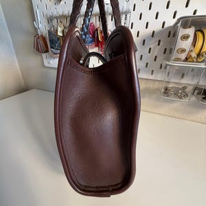May include: A brown leather tote bag with braided handles. The bag has a structured shape and a smooth, textured surface. The interior is not visible. The bag is held up by a person.