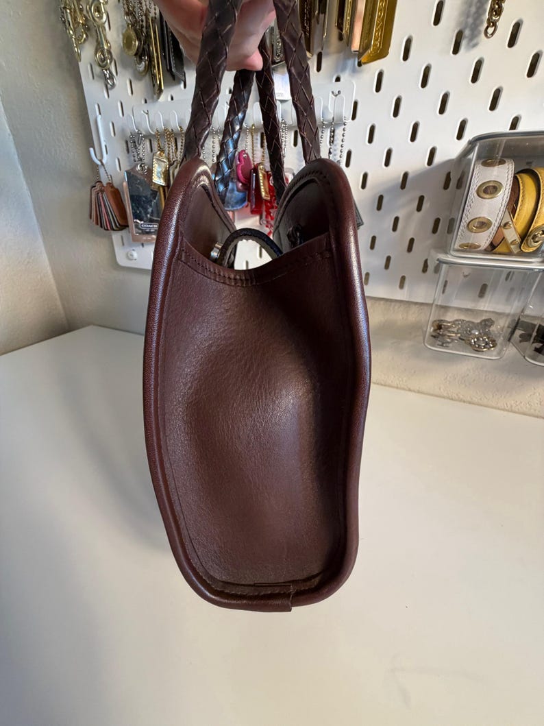 May include: A brown leather tote bag with braided handles. The bag has a structured shape and a smooth, textured surface. The interior is not visible. The bag is held up by a person.