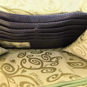 Puede incluir: Vista abierta de una cartera de cuero marr&oacute;n oscuro, que revela m&uacute;ltiples ranuras para tarjetas y un forro interior estampado. El forro presenta un dise&ntilde;o repetitivo de espirales y el nombre de la marca. La cartera est&aacute; abierta, mostrando su organizaci&oacute;n interna.