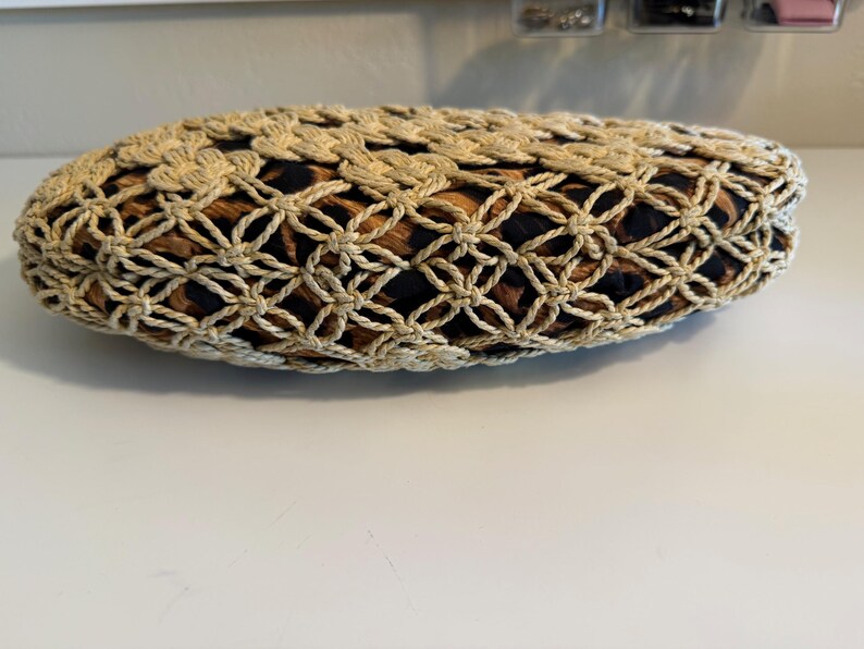 Op de afbeelding: Een beige, ovale clutch met een geweven, opengewerkt ontwerp. De binnenstof heeft een bruine en zwarte dierenprint. De tas ligt op een wit oppervlak.