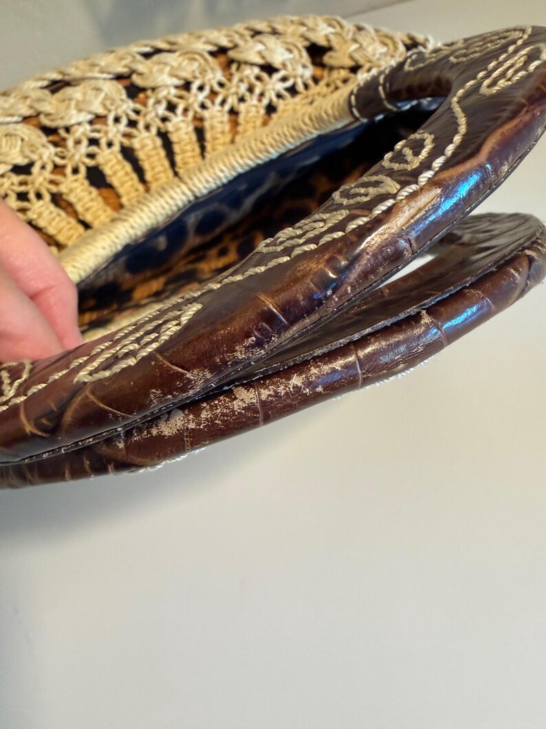 Op de afbeelding: Een vintage handtas met een geweven stro en haakontwerp. De tas heeft een bruin lederen handvat met witte stiksels. De binnenvoering heeft een luipaardprint. De tas is open en onthult de binnenkant.