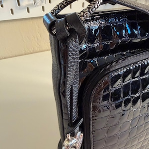 Puede incluir: Bolso de mano de charol negro con un patr&oacute;n en relieve similar al de un cocodrilo. El bolso presenta un asa plateada con un dise&ntilde;o ornamentado y un charm a juego. Una cremallera es visible en la parte delantera.