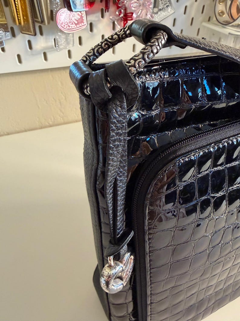 Puede incluir: Bolso de mano de charol negro con un patr&oacute;n en relieve similar al de un cocodrilo. El bolso presenta un asa plateada con un dise&ntilde;o ornamentado y un charm a juego. Una cremallera es visible en la parte delantera.