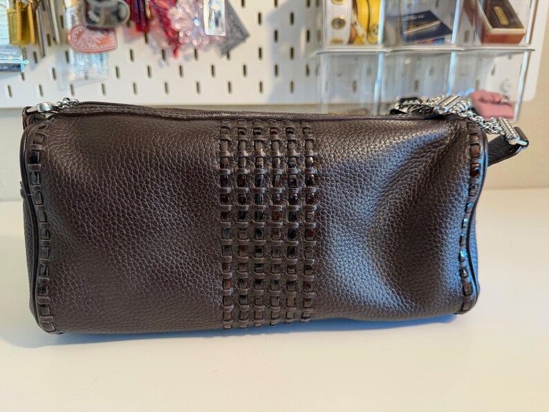 May include: A dark brown leather handbag with a woven center panel. The bag has a rectangular shape with a zipper closure and a silver chain strap. The leather has a textured appearance.