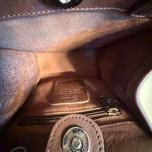 May include: Interior view of a brown leather handbag. The inside features a zippered pocket, a small leather tag, and a magnetic closure. The bag's interior is a lighter shade of brown than the exterior.