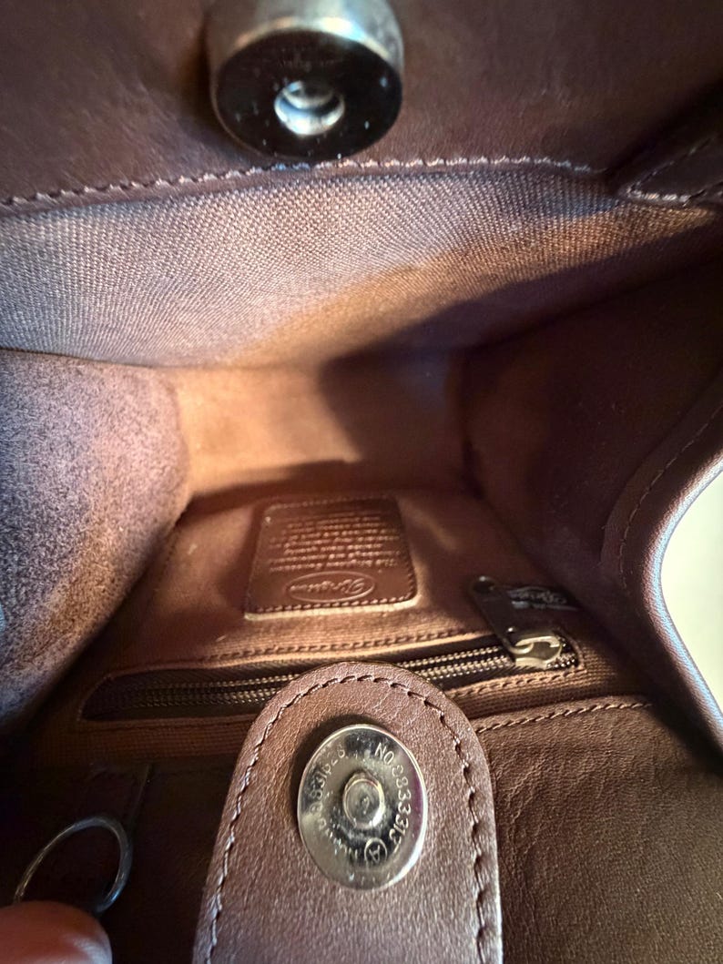 May include: Interior view of a brown leather handbag. The inside features a zippered pocket, a small leather tag, and a magnetic closure. The bag's interior is a lighter shade of brown than the exterior.
