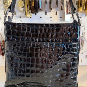 Puede incluir: Bolso de hombro negro brillante con textura de piel de cocodrilo. El bolso tiene forma rectangular con un bolsillo frontal y una correa negra. Cuelga de un panel perforado blanco con varios colgantes.