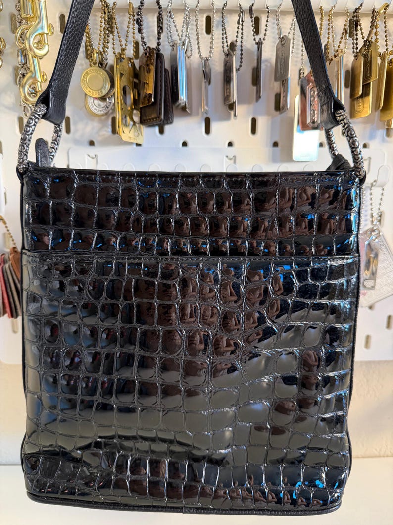 Puede incluir: Bolso de hombro negro brillante con textura de piel de cocodrilo. El bolso tiene forma rectangular con un bolsillo frontal y una correa negra. Cuelga de un panel perforado blanco con varios colgantes.