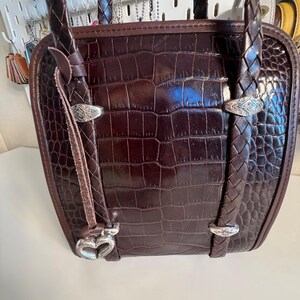 May include: A brown leather handbag with a crocodile skin pattern. The bag has braided handles and silver-toned hardware. A heart-shaped charm hangs from the front.