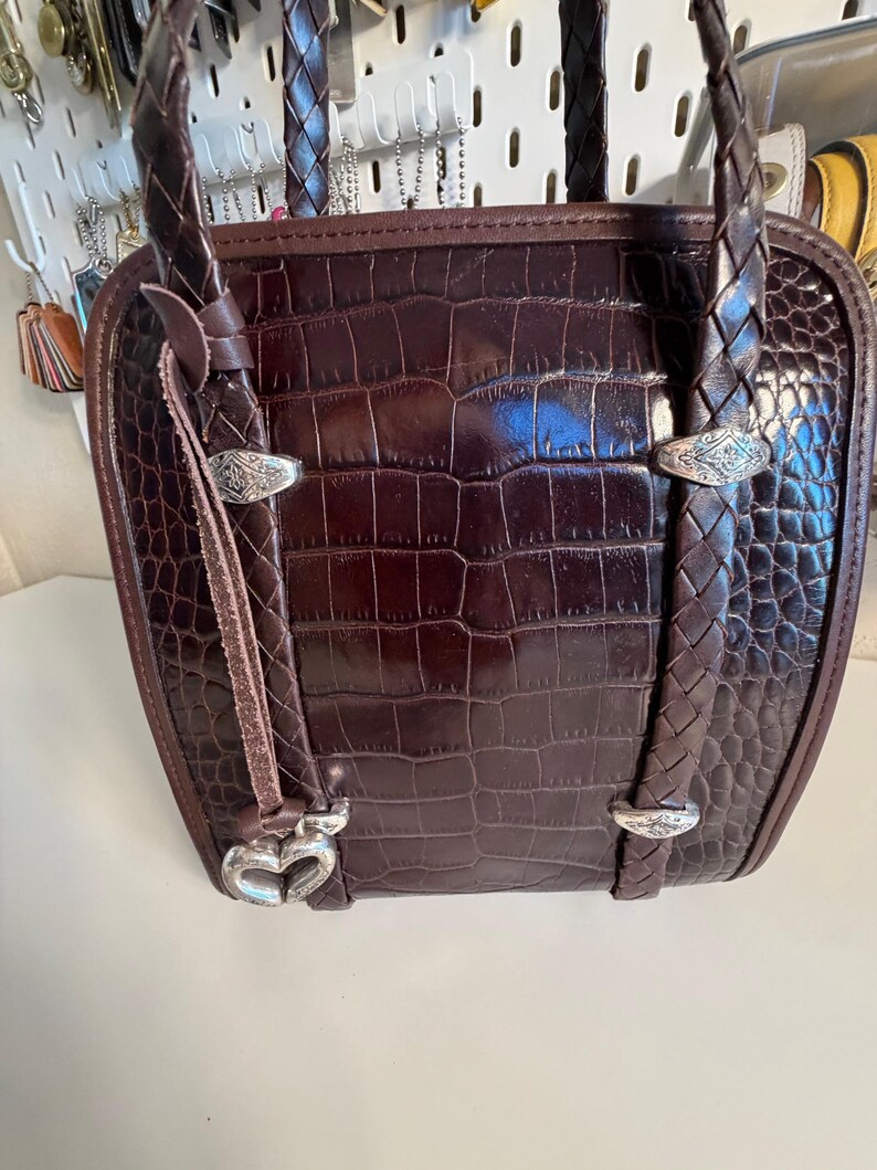 May include: A brown leather handbag with a crocodile skin pattern. The bag has braided handles and silver-toned hardware. A heart-shaped charm hangs from the front.