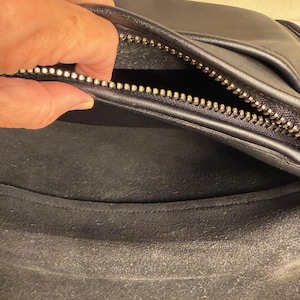May include: Black leather bag with a silver zipper. The interior of the bag is visible, showing a dark, textured lining. The zipper is open, revealing the inside of the bag. The bag appears to be well-worn.