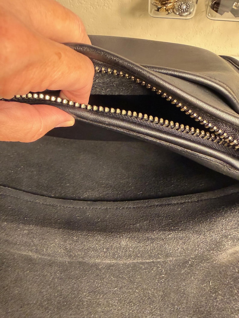 May include: Black leather bag with a silver zipper. The interior of the bag is visible, showing a dark, textured lining. The zipper is open, revealing the inside of the bag. The bag appears to be well-worn.