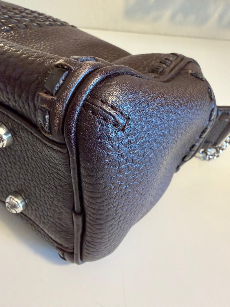 May include: Close-up of a dark brown leather handbag. The bag features a textured surface, visible stitching, and silver-toned hardware. The corner of the bag is visible, showcasing the craftsmanship and design details.