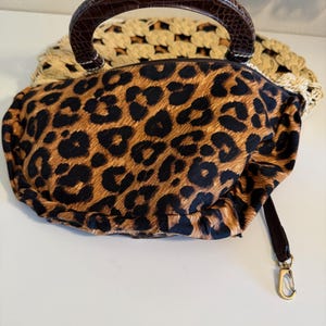Op de afbeelding: Een handtas met luipaardprint met een bruine handgreep en een afneembare riem. De tas heeft een goudkleurige sluiting en een geweven, beige basis. De handgreep heeft een getextureerde, donkerbruine uitstraling.
