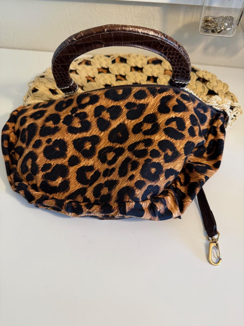 Op de afbeelding: Een handtas met luipaardprint met een bruine handgreep en een afneembare riem. De tas heeft een goudkleurige sluiting en een geweven, beige basis. De handgreep heeft een getextureerde, donkerbruine uitstraling.