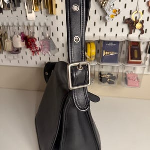 May include: Black leather shoulder bag with a flap closure and silver-tone buckle. The bag has a long, adjustable strap with metal eyelets. The bag's design is classic and minimalist, suitable for everyday use.