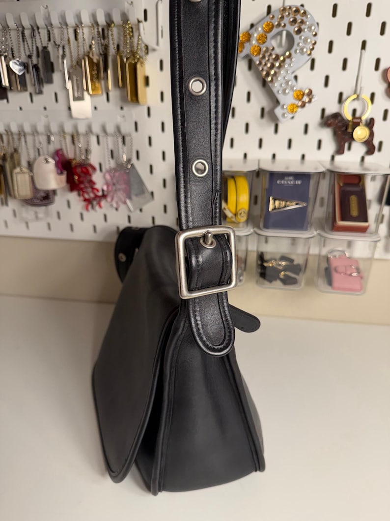 May include: Black leather shoulder bag with a flap closure and silver-tone buckle. The bag has a long, adjustable strap with metal eyelets. The bag's design is classic and minimalist, suitable for everyday use.