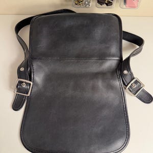 May include: Black leather crossbody bag with a flap closure and adjustable shoulder strap. The bag features silver-tone metal buckles and hardware. The interior is open, and the bag appears to be in good condition.