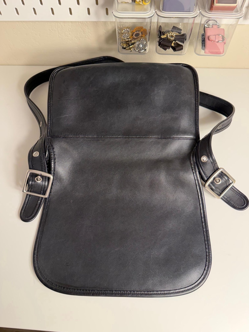 May include: Black leather crossbody bag with a flap closure and adjustable shoulder strap. The bag features silver-tone metal buckles and hardware. The interior is open, and the bag appears to be in good condition.