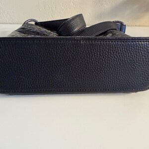 Puede incluir: Primer plano de un bolso de mano de cuero negro. El bolso tiene una superficie texturizada y una base resistente. Las asas son visibles en la parte superior, y el bolso parece estar bien construido y ser duradero. El bolso es de color azul marino oscuro.