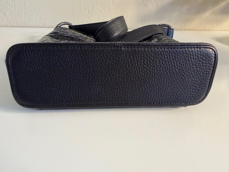 Puede incluir: Primer plano de un bolso de mano de cuero negro. El bolso tiene una superficie texturizada y una base resistente. Las asas son visibles en la parte superior, y el bolso parece estar bien construido y ser duradero. El bolso es de color azul marino oscuro.