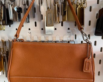 Peut inclure: Un petit sac à main en cuir marron clair avec une poignée supérieure et une fermeture éclair. Le sac présente des coutures blanches et une petite étiquette de marque. Il est suspendu à une étagère blanche avec divers accessoires.