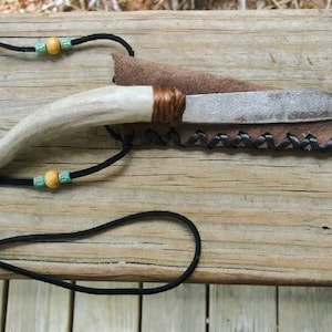 May include: Handmade knife with a bone handle and a leather sheath. The blade is silver, and the handle is wrapped with brown material. A black cord with wooden and turquoise beads is attached.