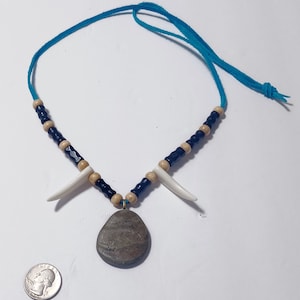May include: A handcrafted necklace featuring a blue suede cord, black and tan beads, and white horn-shaped pendants. The focal point is a gray, teardrop-shaped stone pendant. A U.S. quarter is included for size reference.