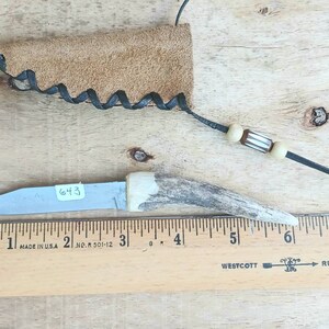 May include: A small, handmade knife with a blade and an antler handle. The knife is accompanied by a leather sheath and a beaded lanyard. A ruler with inch measurements is next to the knife. The blade has the number "643" on it.