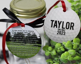May include: A white golf ball ornament with black text that reads "TAYLOR CHS GOLF 2023". The ornament is tied with a red ribbon and is sitting on a green plant.